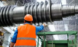 Pracownik w pomarańczowej kamizelce i kasku wskazuje na ogromny wał turbiny. To przykład precyzyjnej produkcji jednostkowej.