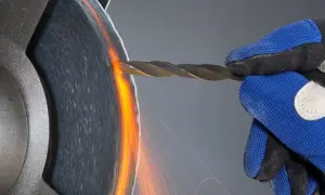 Ręka w niebieskiej rękawicy szlifuje wiertło, tworząc iskry. Precyzyjne wiercenie w aluminium wymaga ostrości.