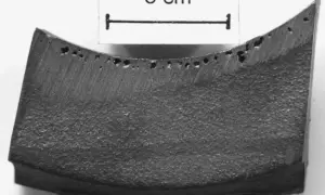 Fragment metalu z licznymi porami, widoczne wady odlewnicze. Skala 3 cm.