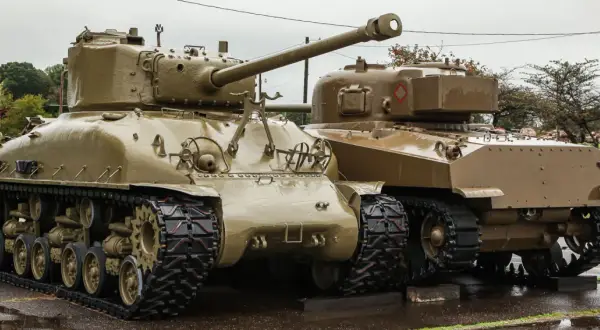 Dwa czołgi Sherman, których kraj pochodzenia to USA, stoją obok siebie na mokrej nawierzchni.
