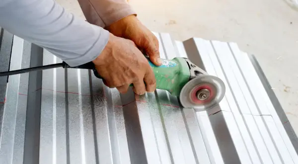 Ręce trzymają szlifierkę kątową, która tnie falistą blachę aluminiową wzdłuż czerwonej linii. To pokazuje, czym ciąć aluminium.