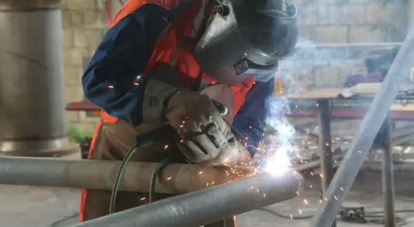 Spawacz w masce i rękawicach łączy metalowe rury. To praca, do której wykorzystuje się spawarkę inwertorową, generującą jasne iskry.
