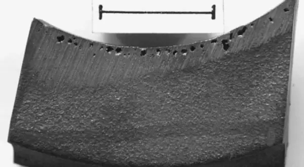 Fragment metalu z licznymi porami, widoczne wady odlewnicze. Skala 3 cm.