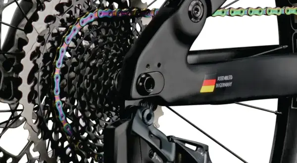 Kolorowy łańcuch rowerowy oplata kasetę zębatek. Widać ramę z napisem "ASSEMBLED IN GERMANY" i flagą. Gwint trójkątny jest kluczowy dla połączeń.