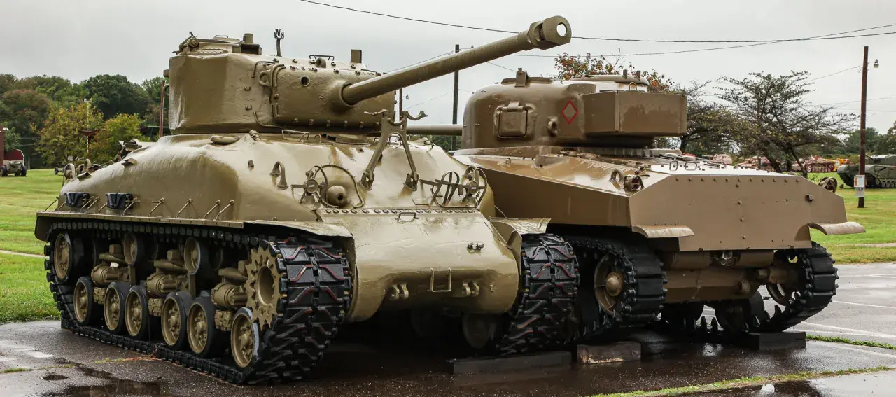 Dwa czołgi Sherman, których kraj pochodzenia to USA, stoją obok siebie na mokrej nawierzchni.