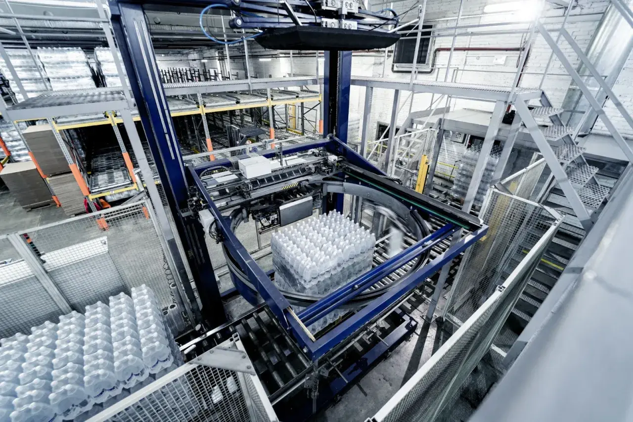 Automatyczne gniazdo produkcyjne pakuje butelki z wodą. Maszyna obraca paletę z napojami, przygotowując ją do dalszego transportu.