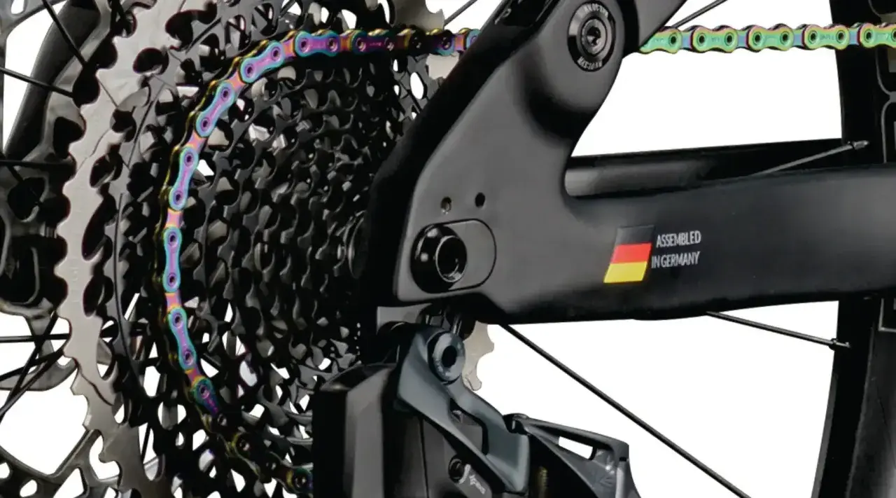 Kolorowy łańcuch rowerowy oplata kasetę zębatek. Widać ramę z napisem "ASSEMBLED IN GERMANY" i flagą. Gwint trójkątny jest kluczowy dla połączeń.