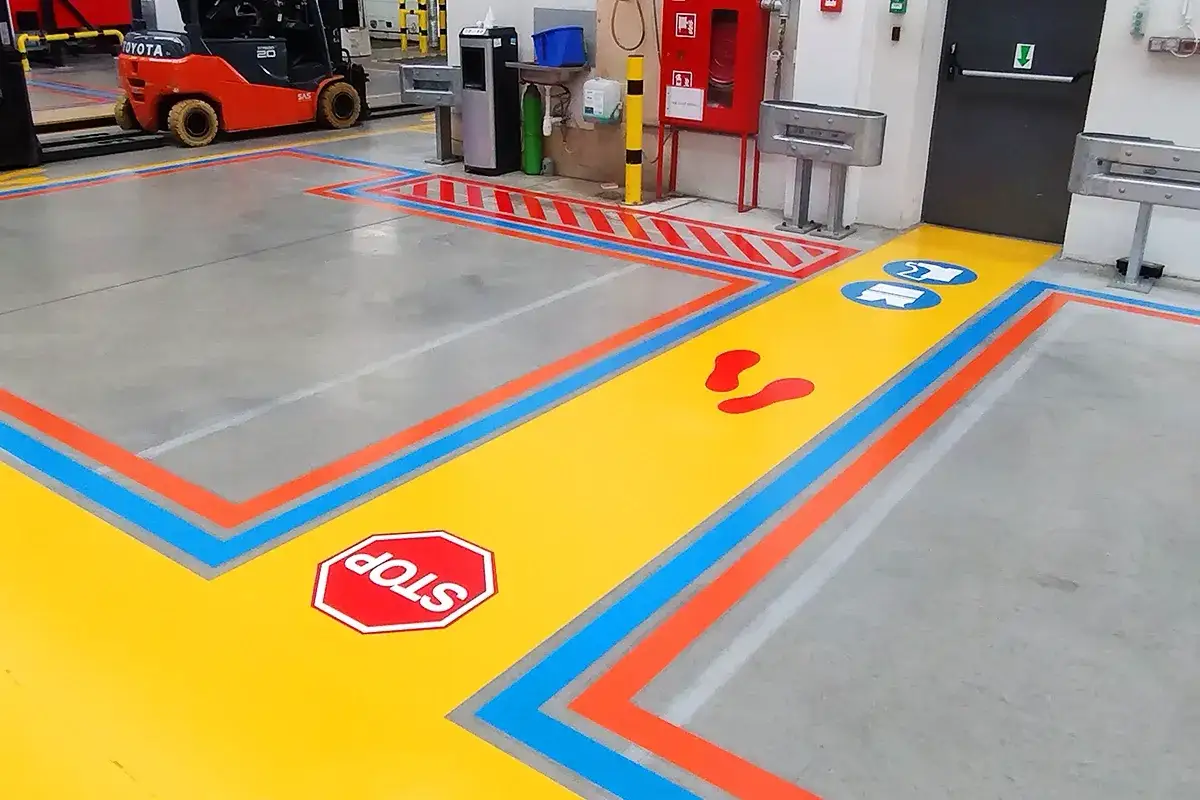 Jasna ścieżka z oznaczeniami, w tym ślady stóp i znak STOP, to przykład metody kaizen wdrożonej na hali produkcyjnej dla bezpieczeństwa.
