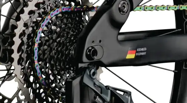 Kolorowy łańcuch rowerowy oplata kasetę zębatek. Widać ramę z napisem "ASSEMBLED IN GERMANY" i flagą. Gwint trójkątny jest kluczowy dla połączeń.